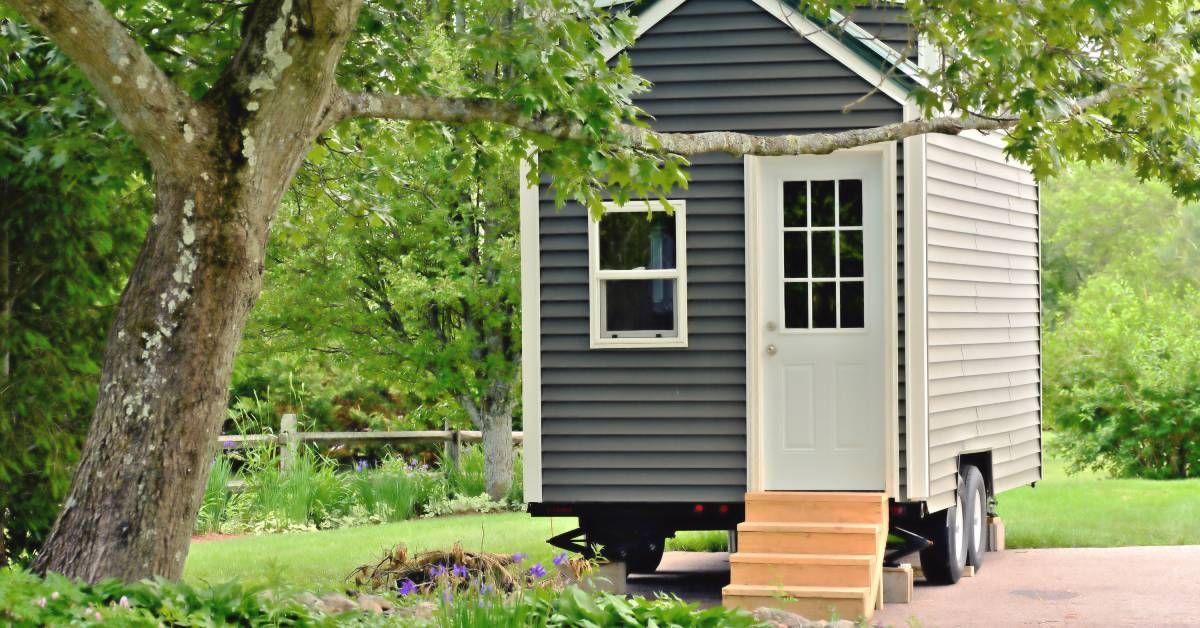 A tiny home with gray siding and a white front door that is surrounded by trees. The home is on wheels.