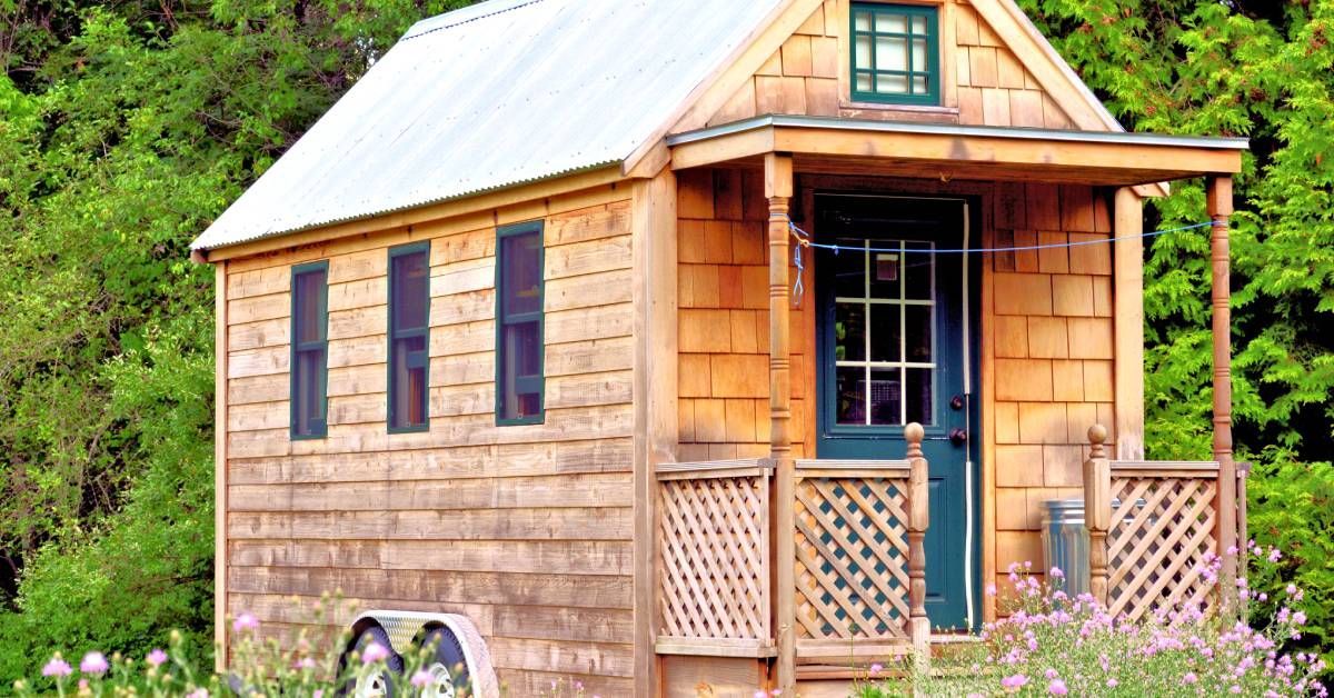 The exterior of a wooden tiny house with a small front porch on wheels. The house is surrounded by trees.