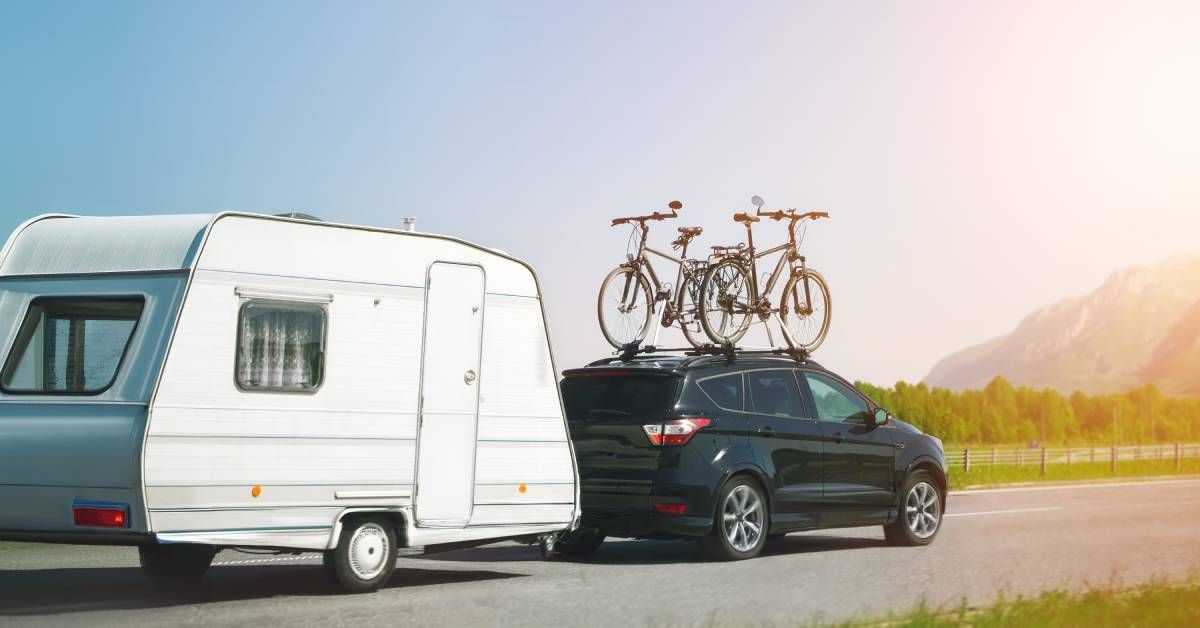 A black SUV with two bikes mounted on the roof is towing a small white RV. There are mountains in the background.