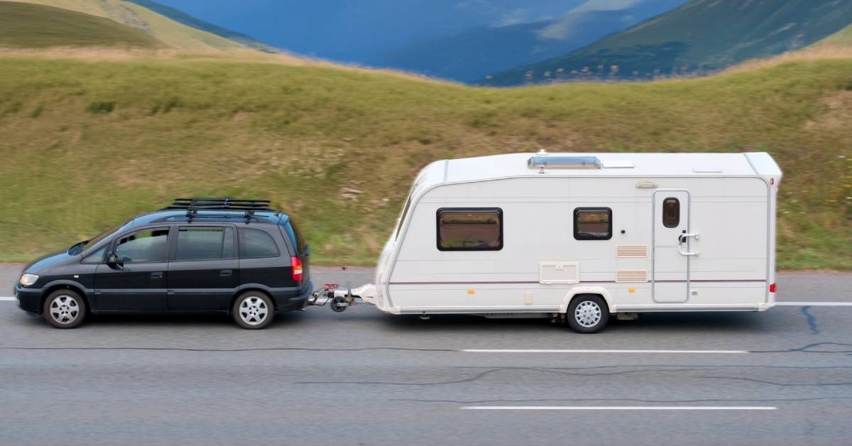 A black car is towing a white RV. There is grass and a large mountain range on the side of the road.