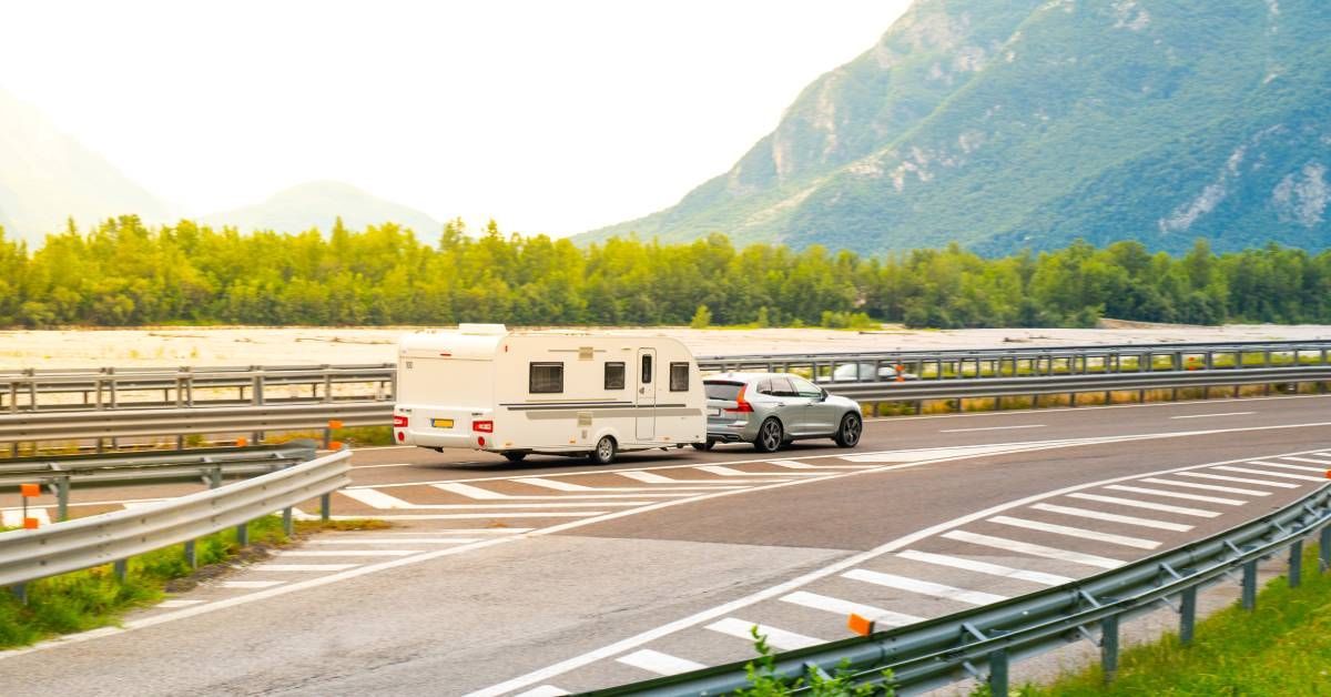 A silver SUV is towing a large white RV. The car is driving on an open road through the mountains on a sunny day.