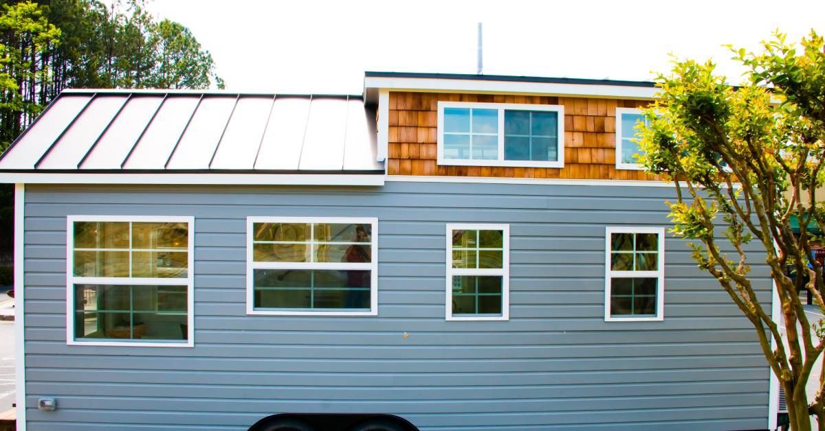 A tiny home with blue siding, white window frames, and a metal roof. The home is set on wheels.