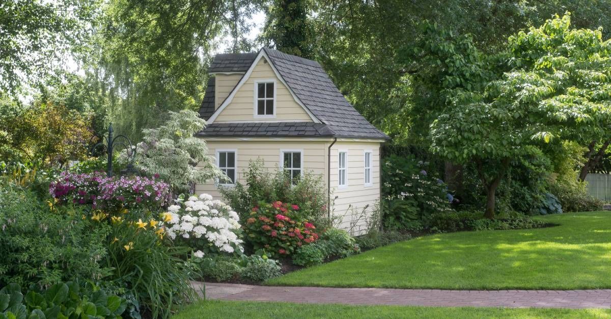 A tiny home with cream siding and a black roof. Lush landscaping surrounds the house.