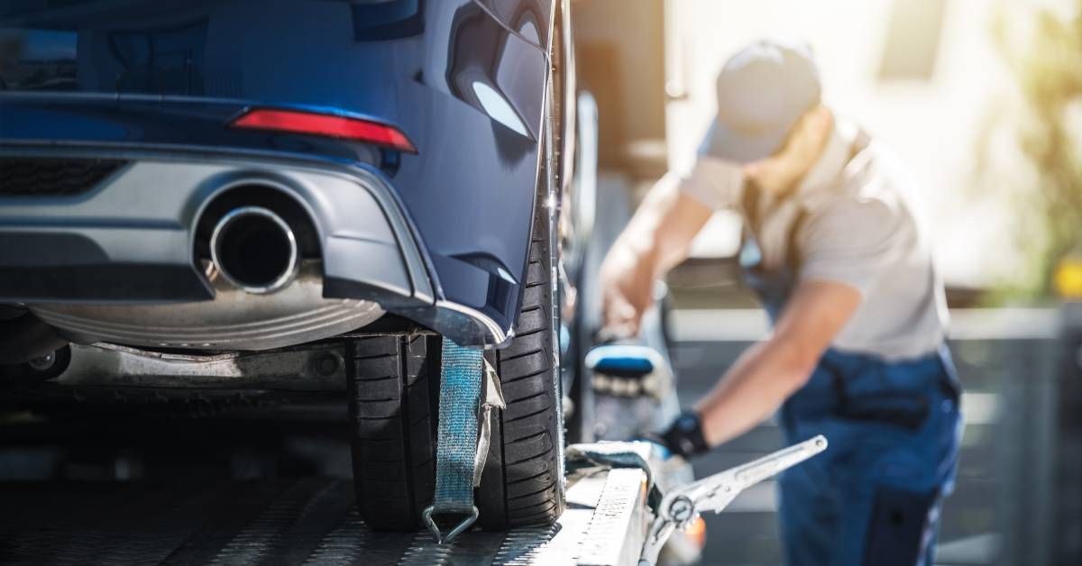 A man dressed in blue overalls and a blue hat using straps to secure a blue vehicle on a trailer.