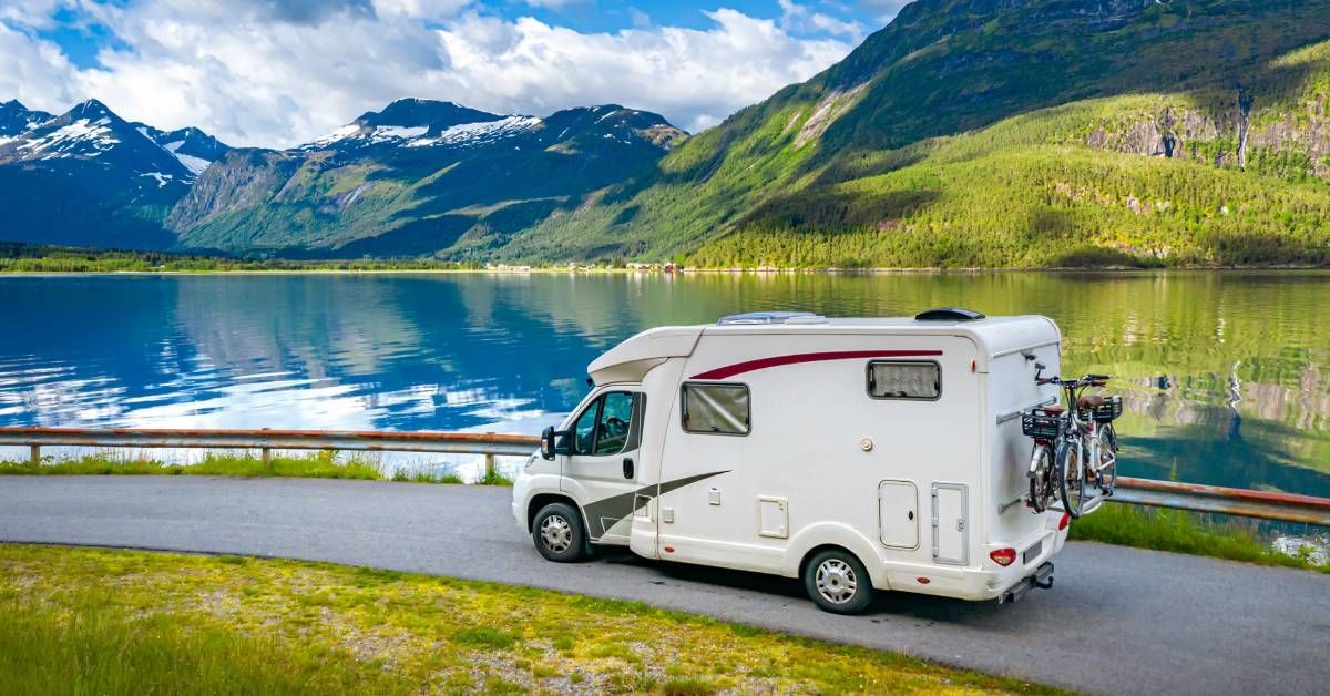 A white camper van with bikes attached to the back is driving down a road alongside a lake and mount