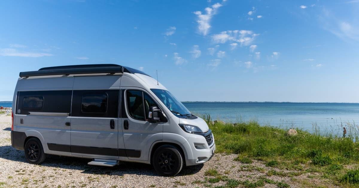 A white camper van parked on gravel next to a body of water. There is tall grass on the shoreline.