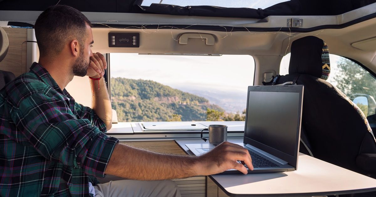 A young man wearing a plaid shirt working on a laptop in an RV while looking out the window at the scenic view.