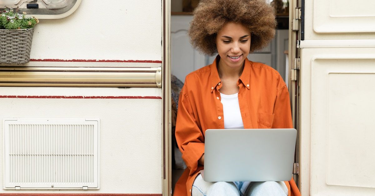 A young woman wearing an orange shirt and jeans working on a laptop while sitting in the doorway of an RV.