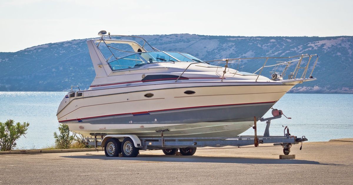 A large white and red boat sitting on a metal trailer with wheels in front of a body of water and hilly landscape.