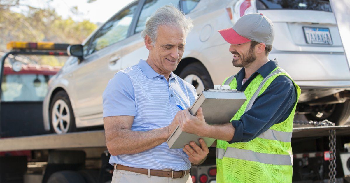 A man in a safety vest holding a clipboard as another man signs it. They're in front of a tow truck.