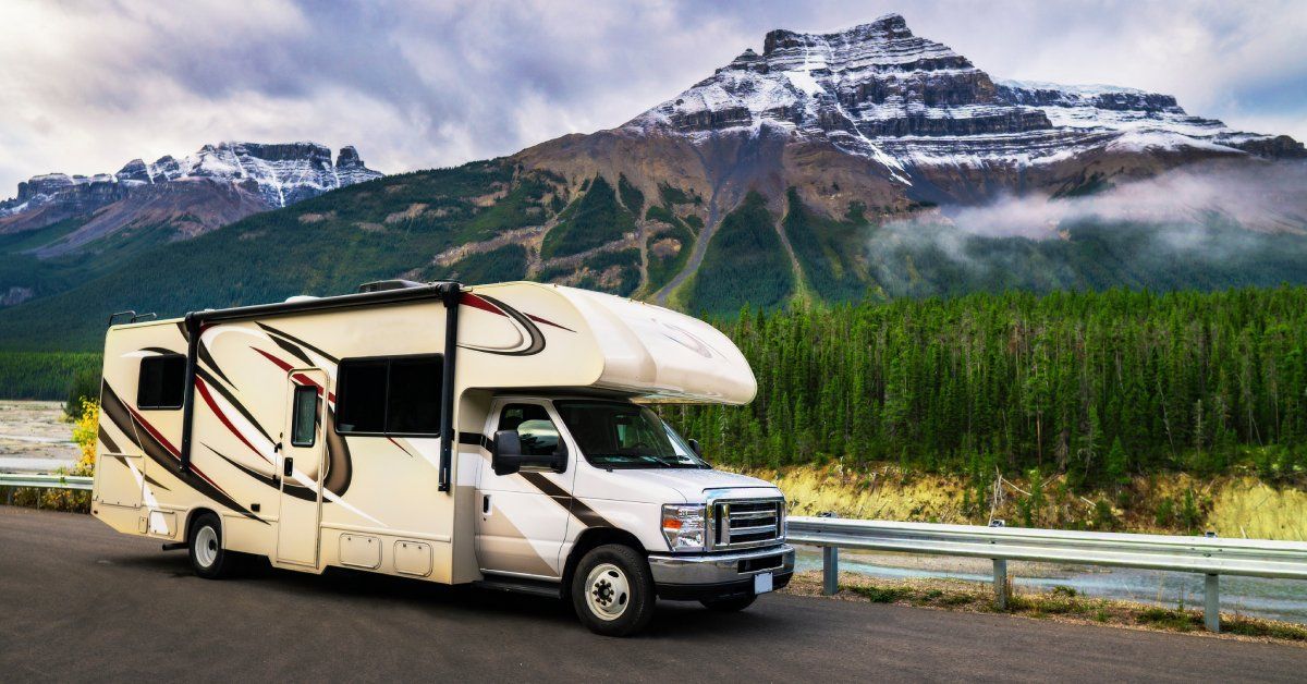 A white RV with red and black accents parked on a road in front of mature trees and large mountains.