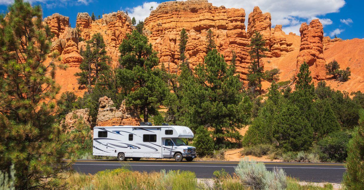 A white RV driving near mature trees and large red rock formations on a sunny day.