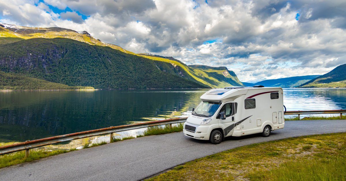 A white RV driving along a large body of water. Mountains and a cloudy sky are in the background.