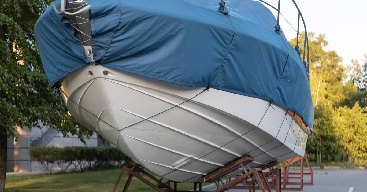 A white motorboat with a blue fabric cover sitting on a wooden stand in a parking lot with trees in the background.