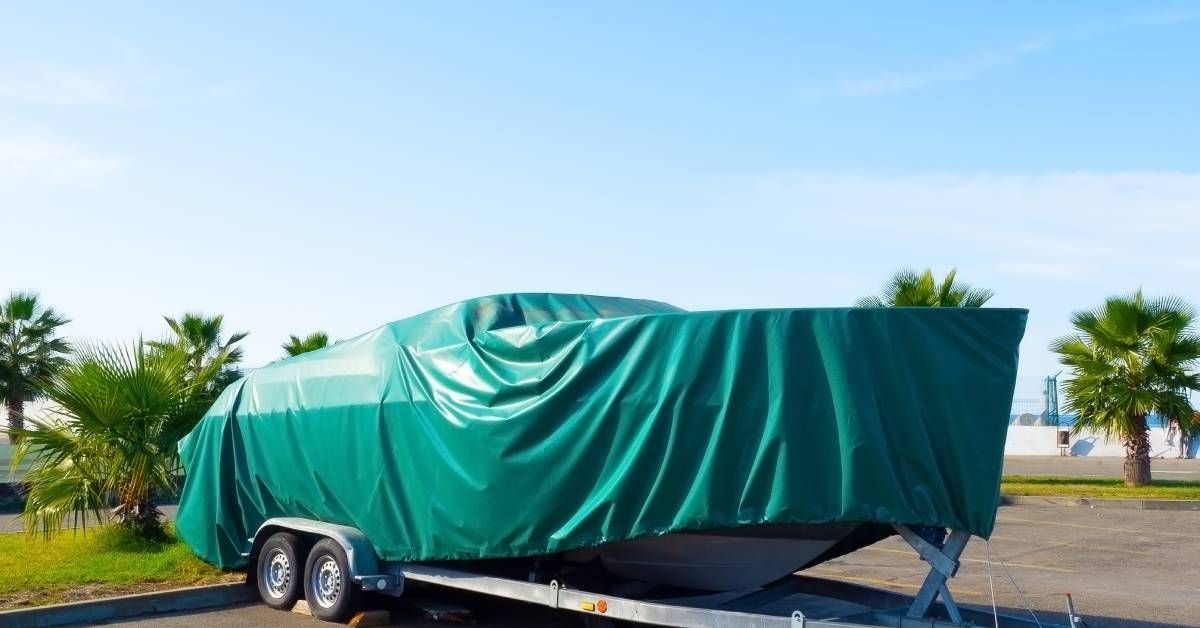 A motorboat covered in a green tarp sitting on a metal trailer with wheels. Short palm trees are in the background.