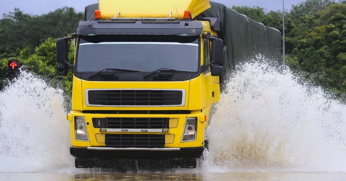 The front of a large truck with a yellow cab making a big splash as it drives through standing water.