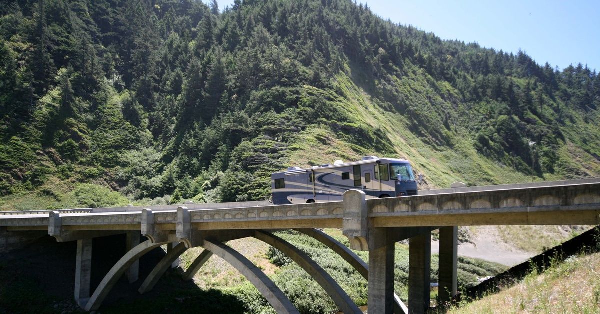 A blue and white RV driving over a cement bridge with large tree-lined hills in the background on a sunny day.