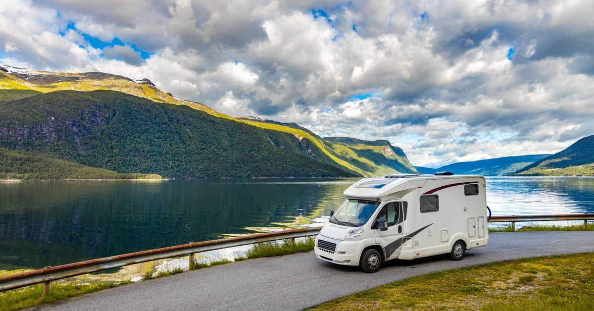 A white RV with red and gray accents driving down a road with a body of water and green hills in the background.
