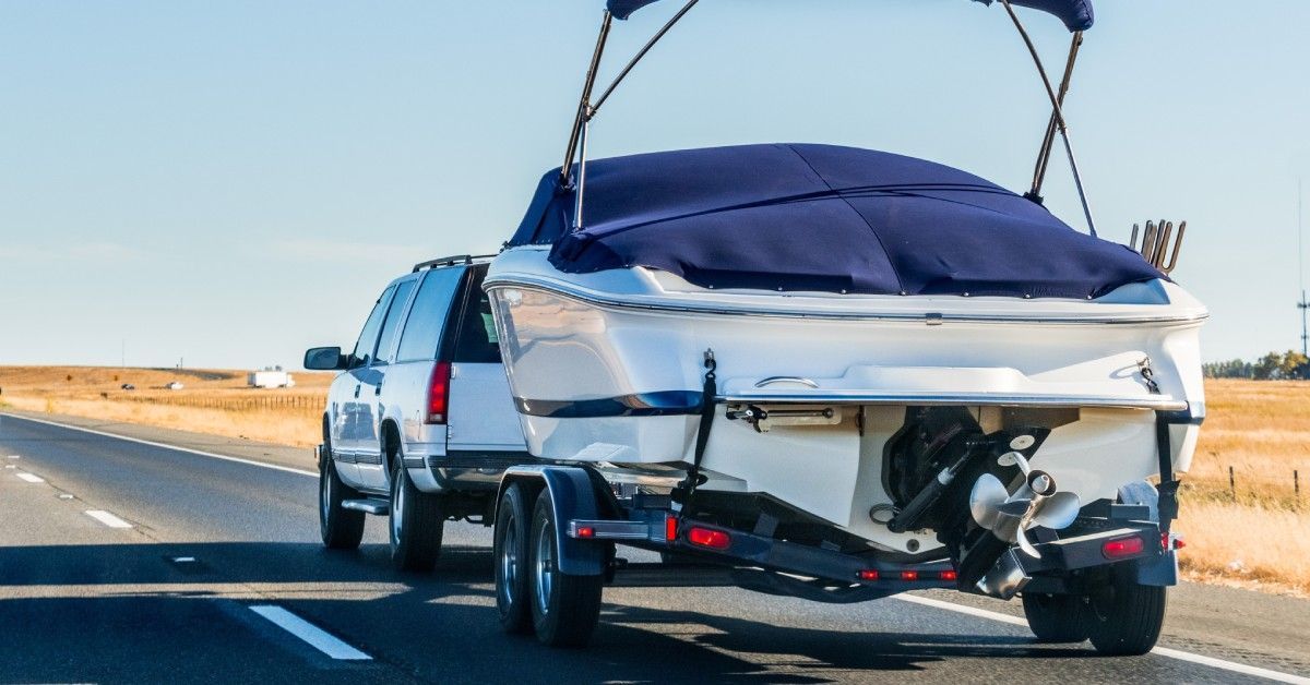 A white SUV towing a white motorboat with a blue cover on an open road next to a field with brown grass.
