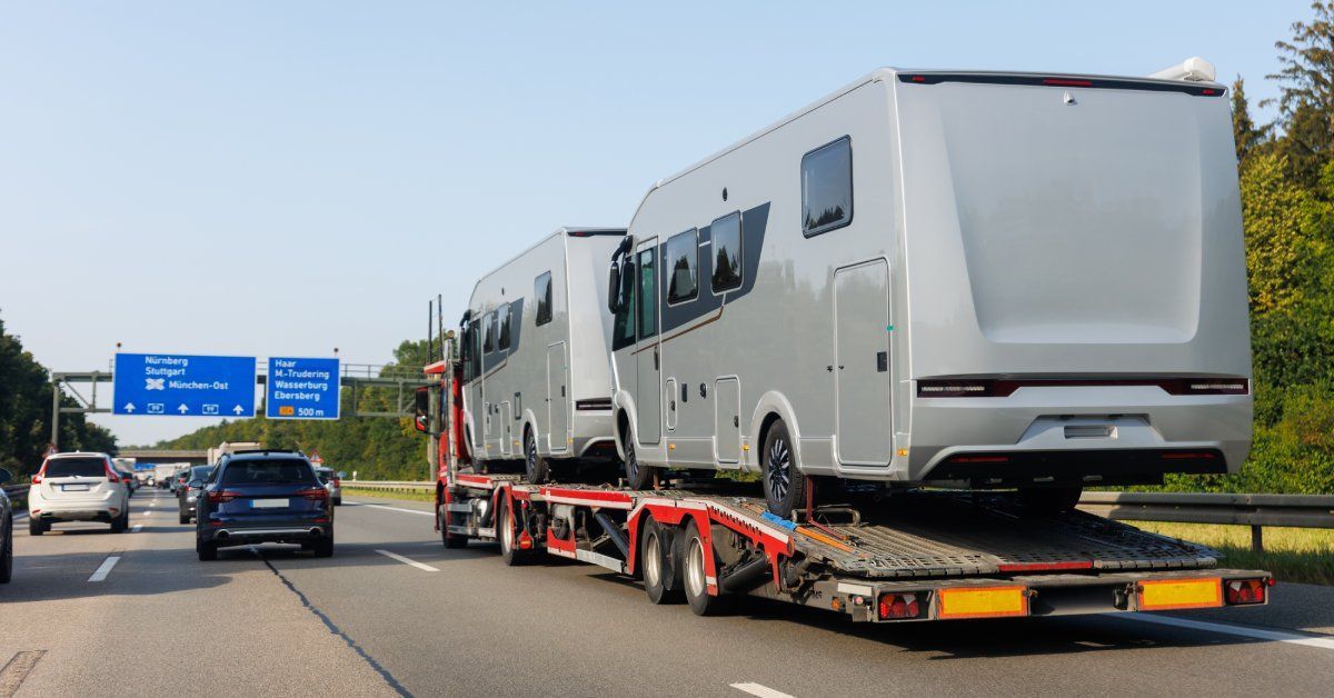 A truck with a red trailer transporting two large gray RVs on a busy, multilane highway.