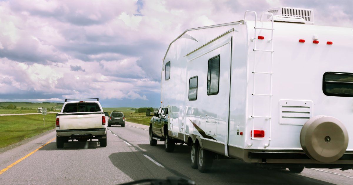 A black pickup truck towing a white RV on a busy two-lane highway surrounded by open fields on a cloudy day.