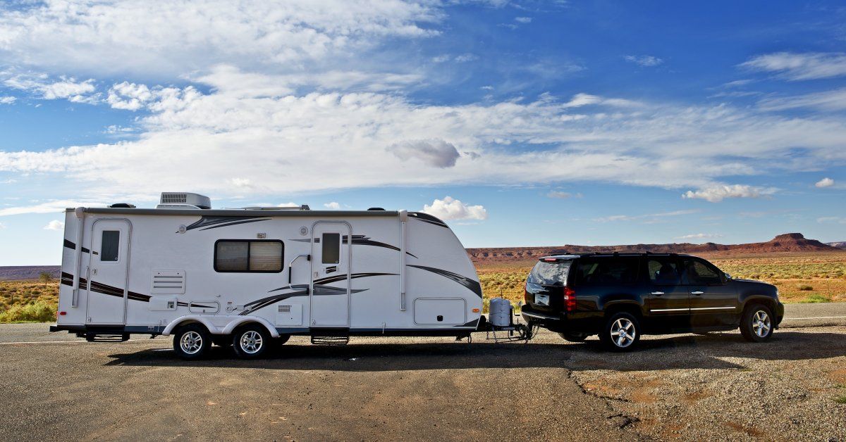 A black SUV towing a large white RV with a trailer hitch drives along a road surrounded by a desert 