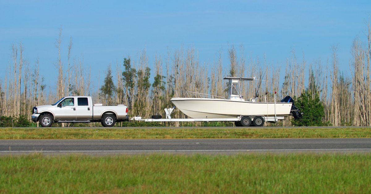 A white pickup truck towing a white fishing boat on a trailer drives along a road flanked by grass and trees.
