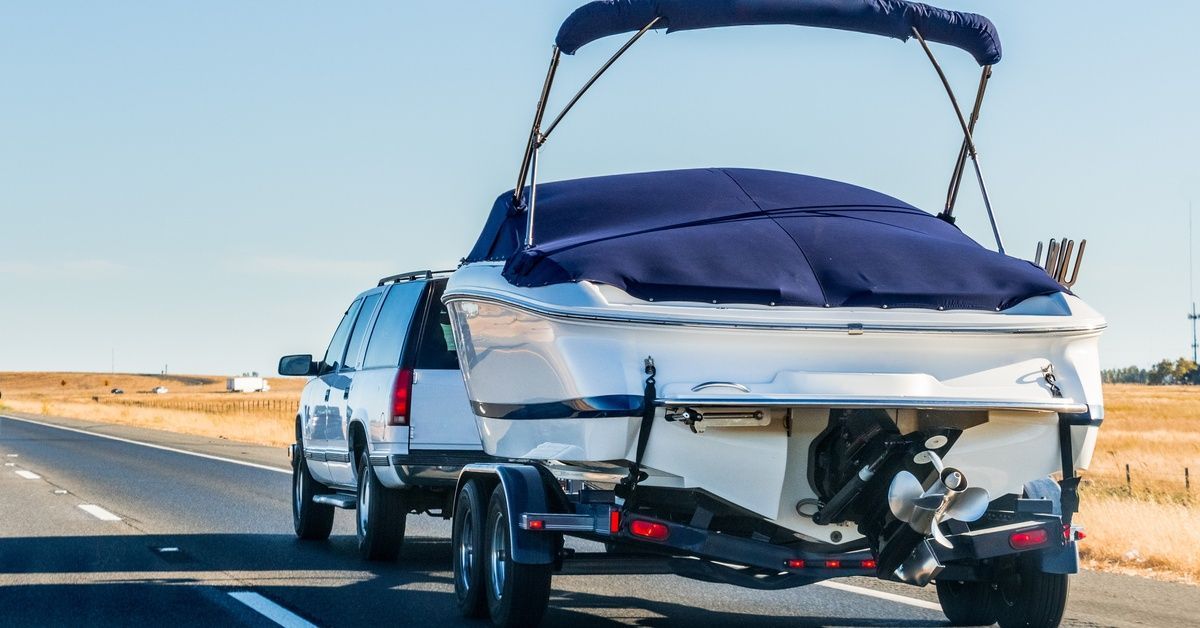 An SUV towing a white boat with a single engine and blue cover on a road surrounded by open fields.