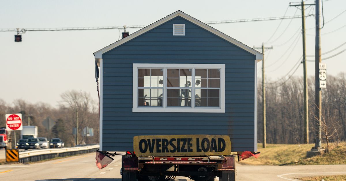 A blue tiny house rests on the back of a trailer on a roadway with a yellow banner labeled 