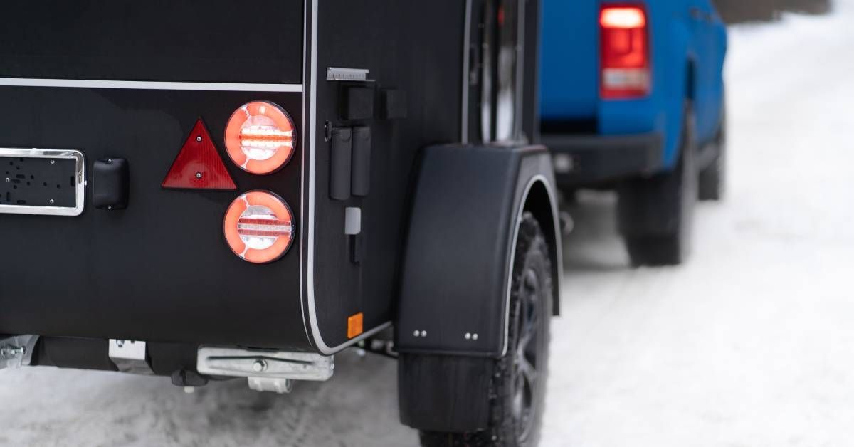 A close-up of the back of a black camper trailer and the back of a blue truck towing it down a snowy