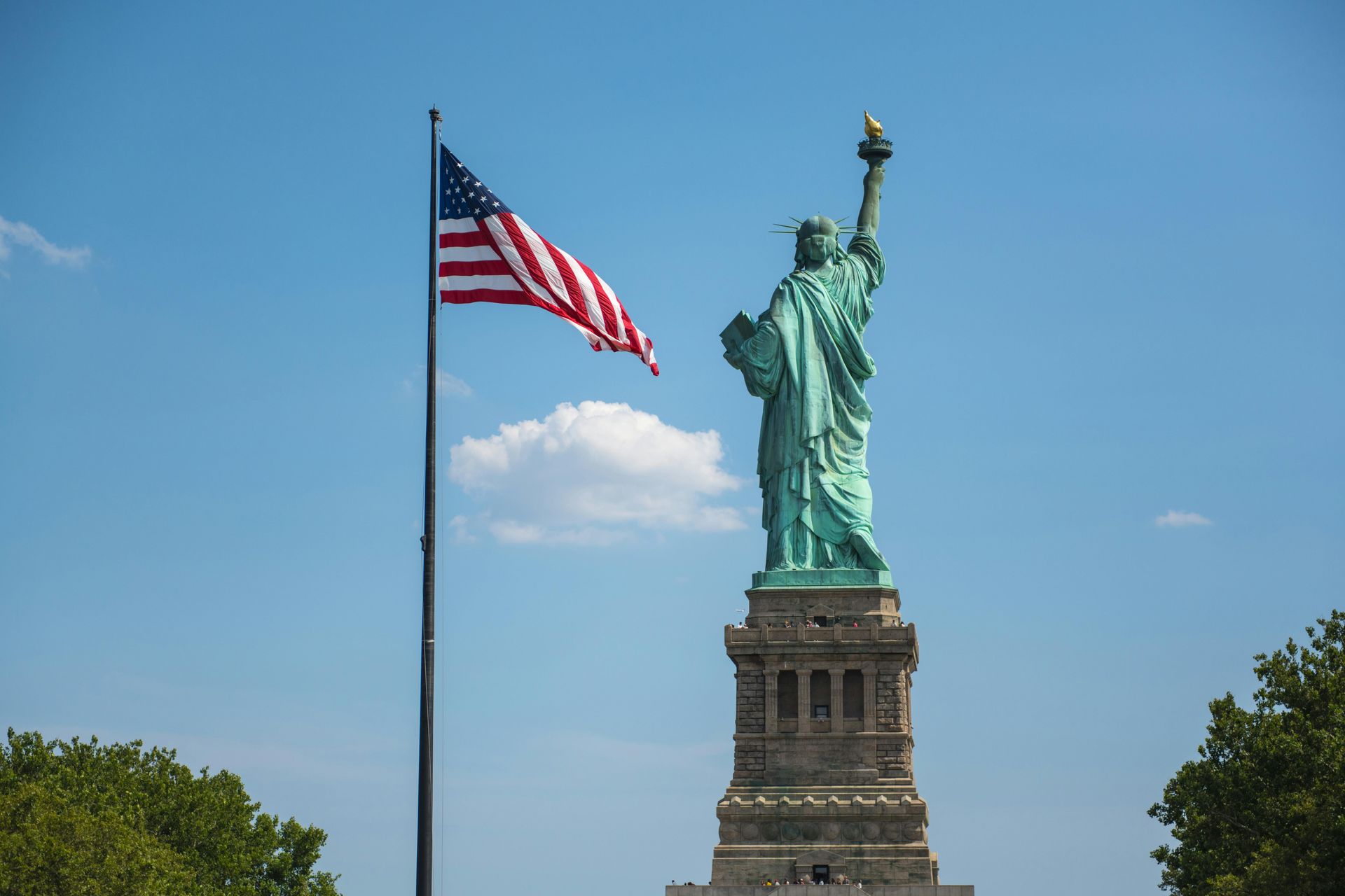 The statue of liberty is surrounded by trees and a flag is flying in front of it.