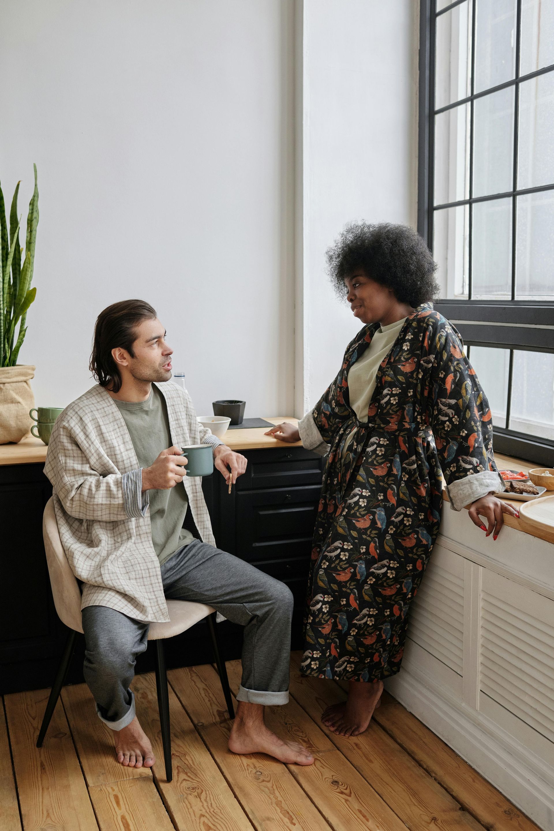 A man is sitting in a chair next to a woman standing next to a window.