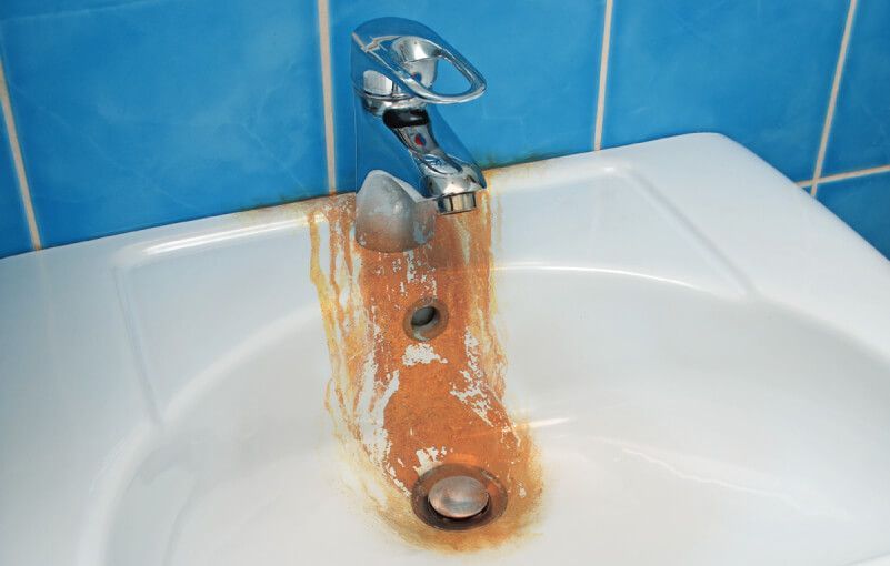 Rusty faucet dripping brown water into a white sink. Blue tile wall background.