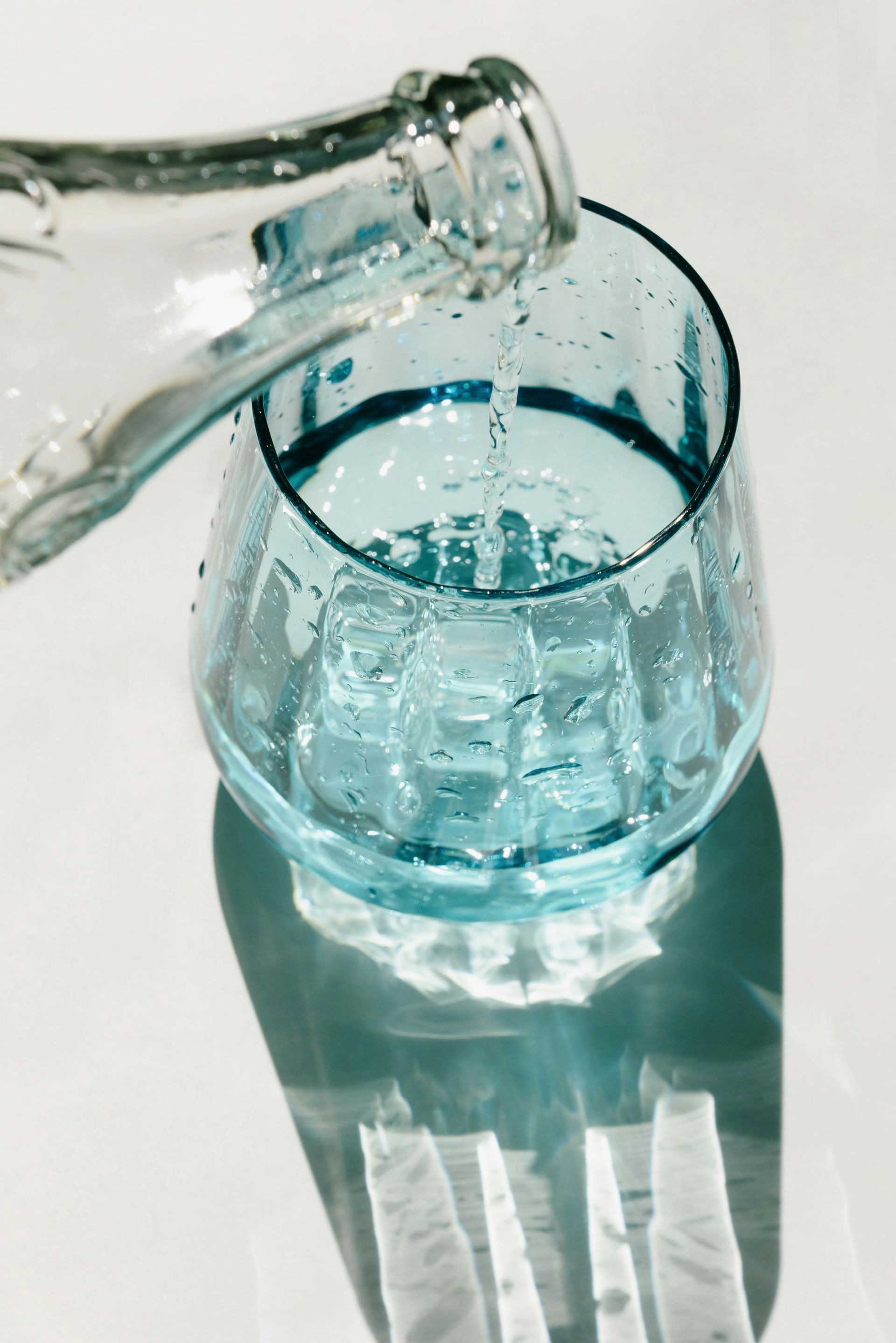 Water pouring from a bottle into a blue glass with ice cubes on a white surface.
