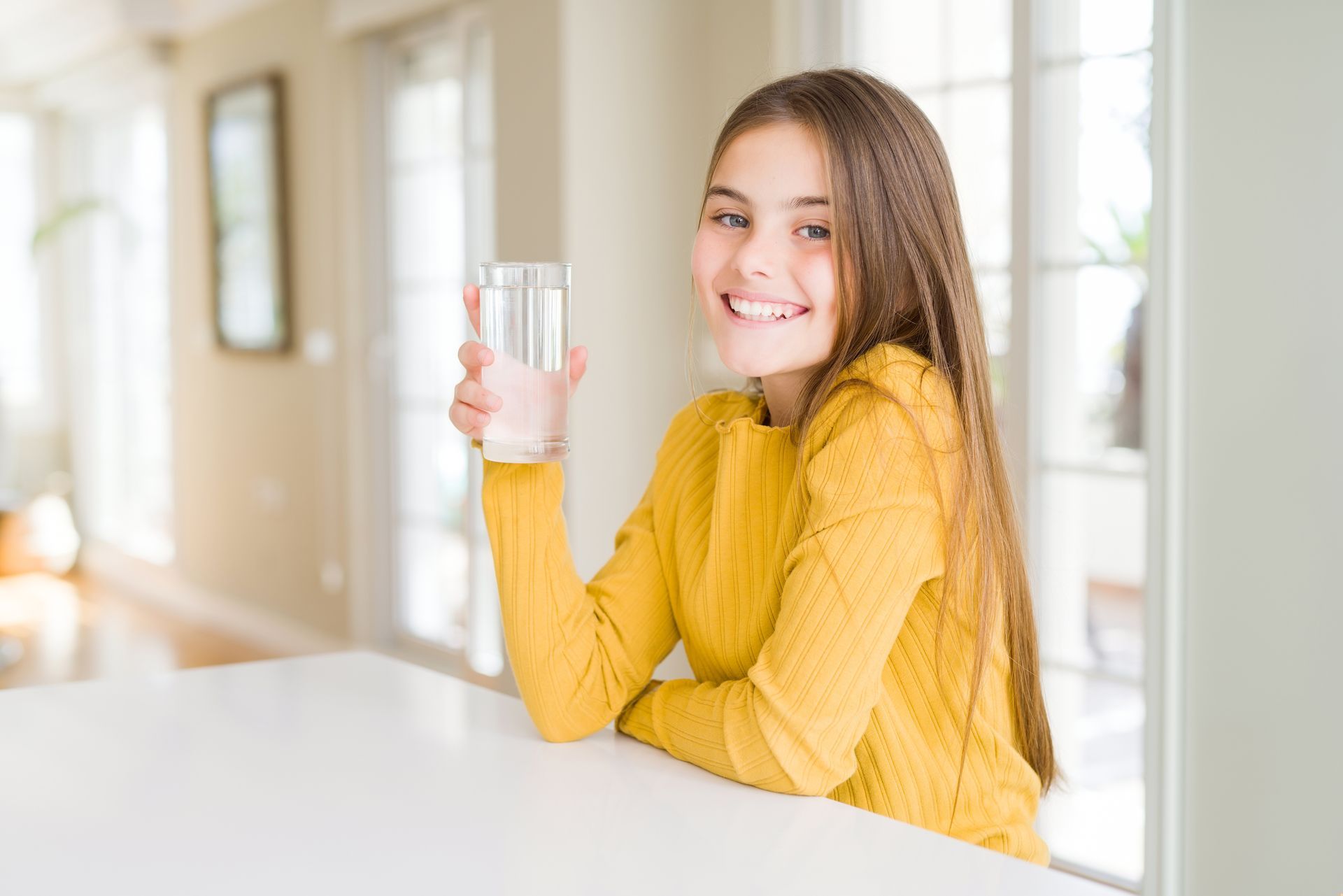Girl with long brown hair smiles, holding a glass of water, seated at a white table.