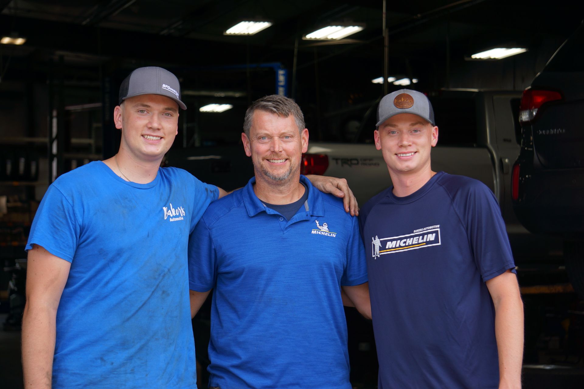 Three men in a garage, two in blue shirts, one in a blue polo, all smiling.