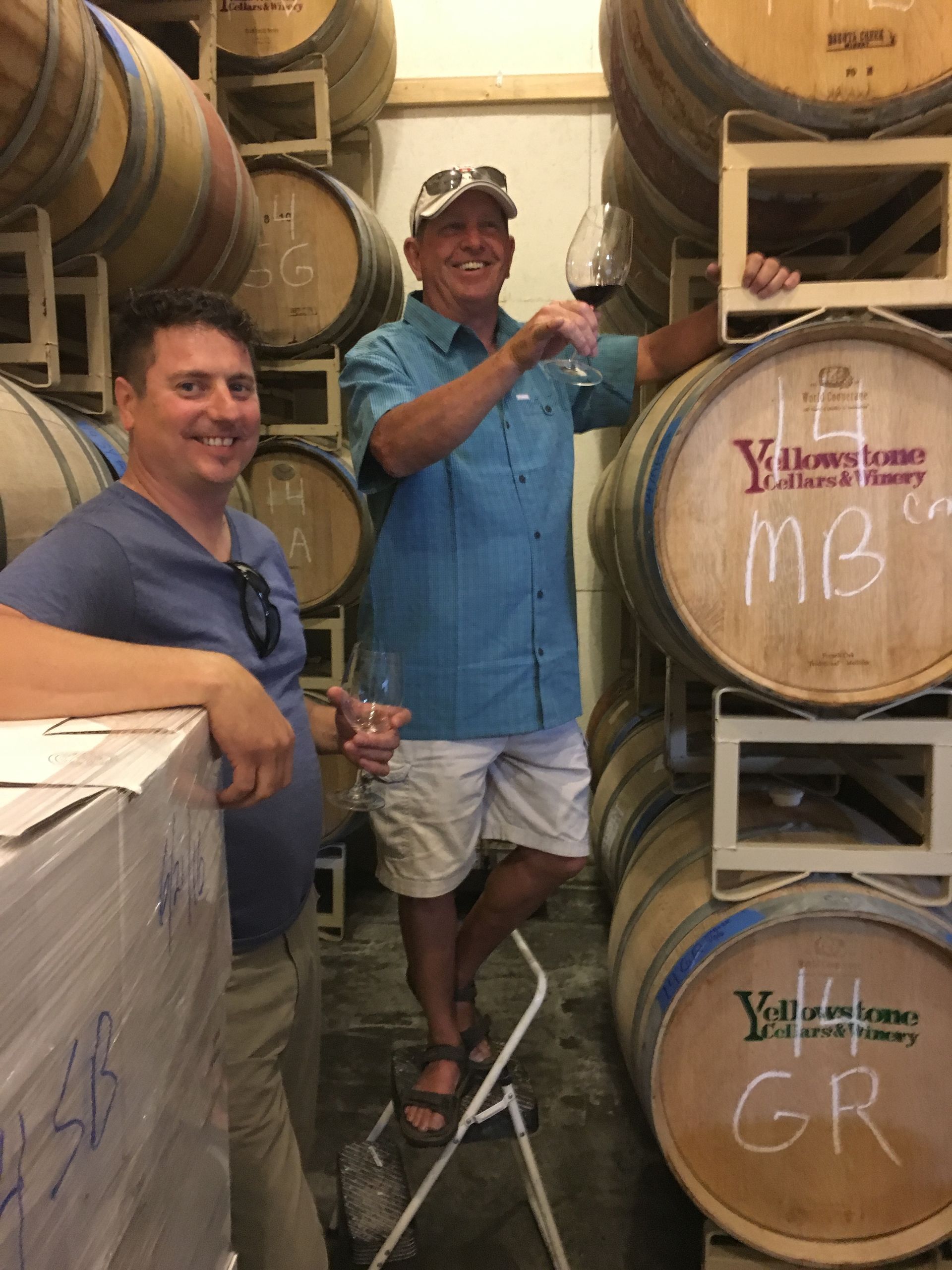 Two men are standing in front of a stack of wine barrels one of which says mb on it