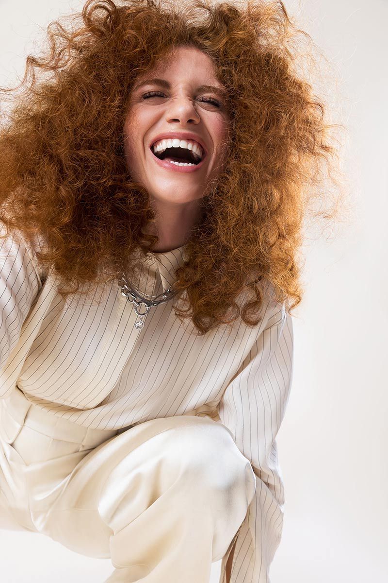 Woman With Red Curly Hair Is Wearing A White Sweater And White Pants And Smiling — Siddha Hair Studio in Currajong, QLD