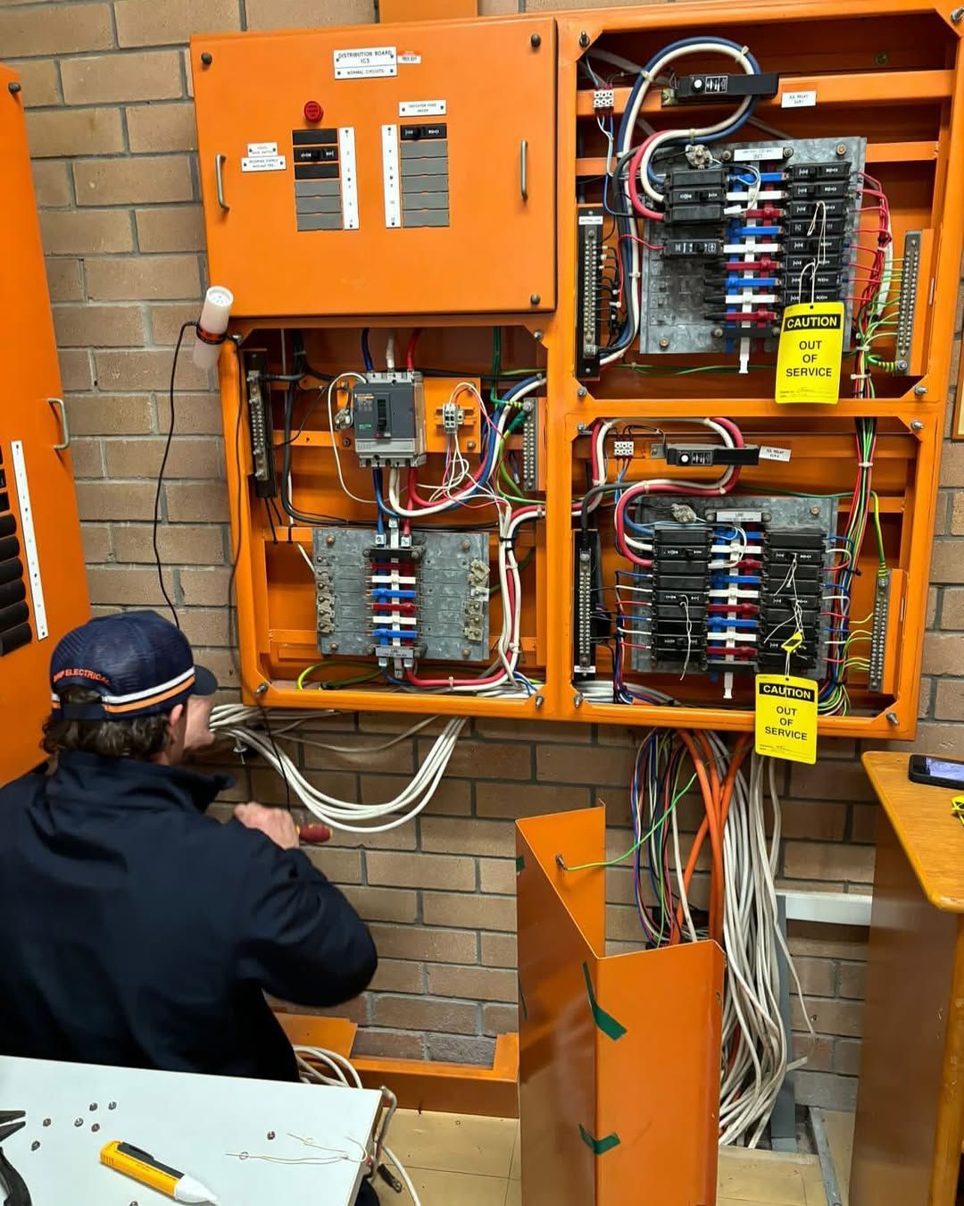 An electrician works on an open orange electrical panel on a brick wall. Wires and components are visible.