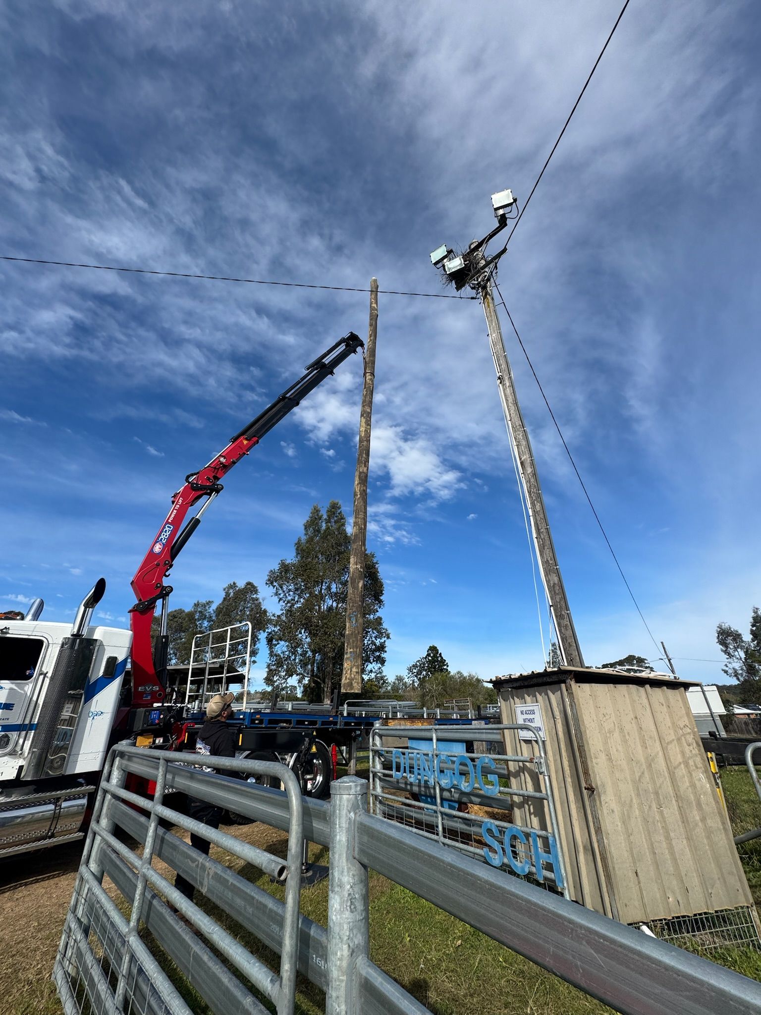 A crane lifts a wooden pole near a utility pole with equipment, fence in foreground, blue sky.