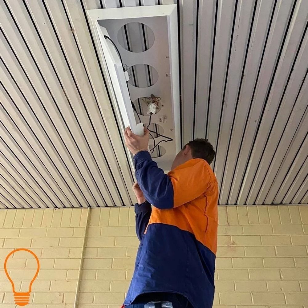 Person on a ladder installing a light fixture in a ceiling. The person wears an orange and blue jacket.