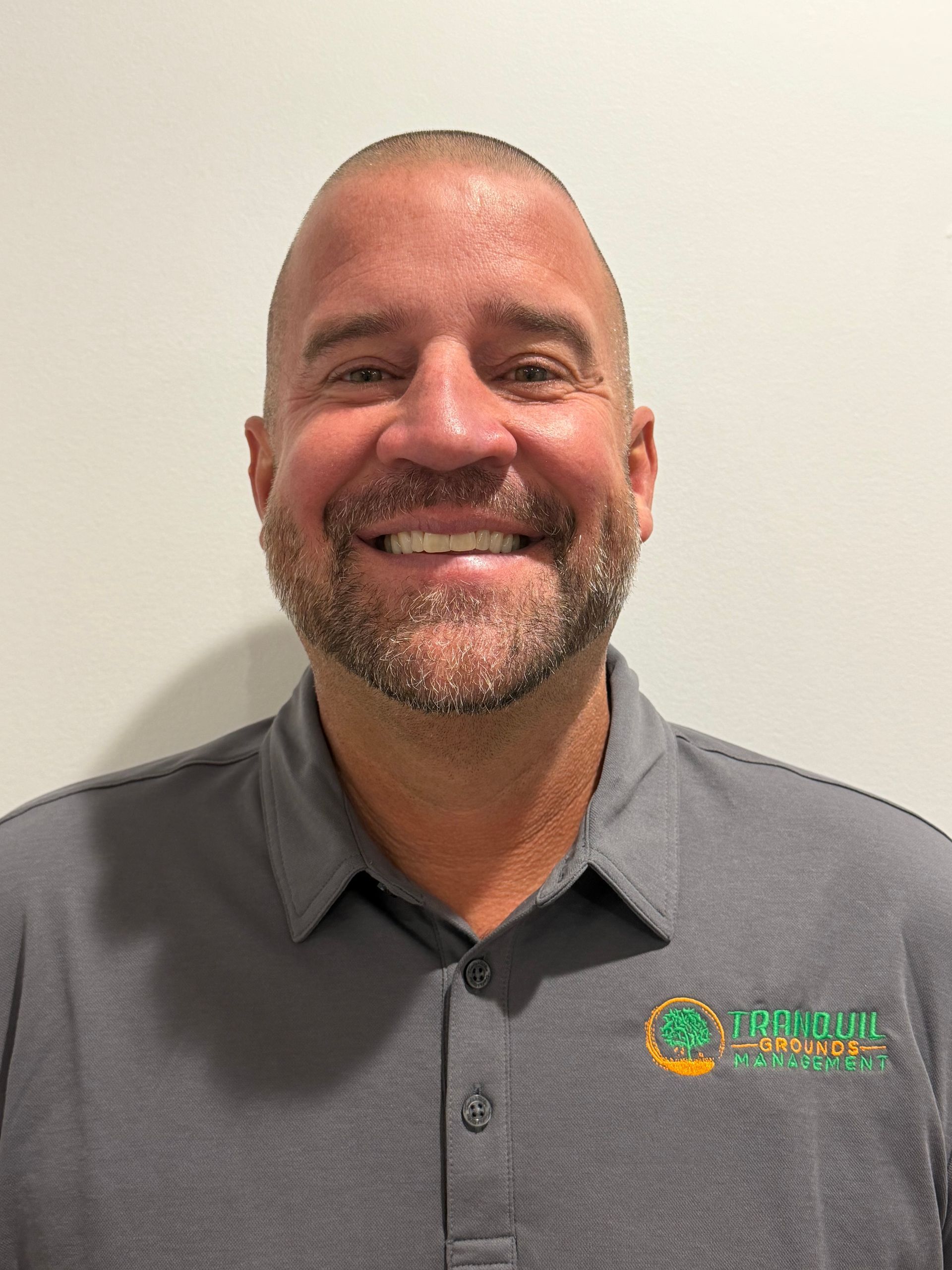 Man with short hair and a beard smiling, wearing a grey polo shirt with a logo.
