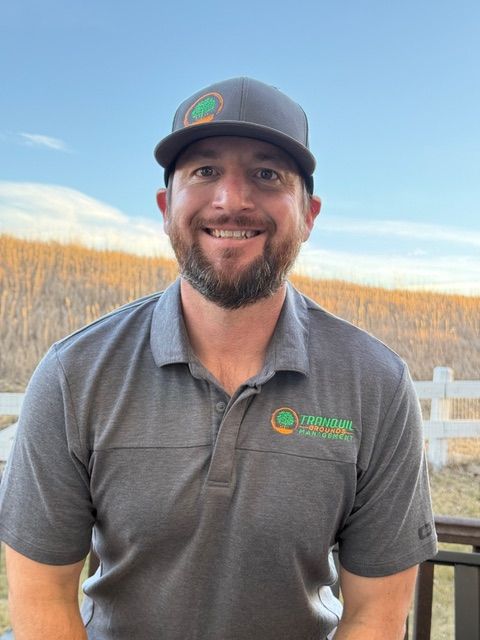 Man in gray cap and shirt smiles outdoors, logo visible.