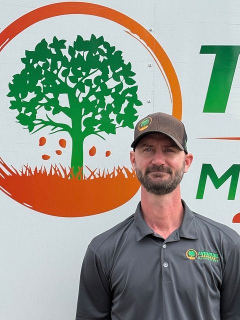 Man in gray polo shirt and cap smiles in front of a company logo with a green tree.