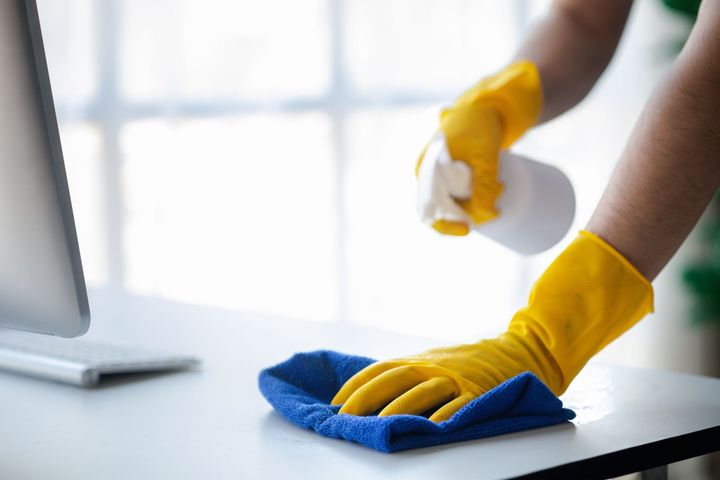 Person in yellow gloves spraying and wiping a white desk with a blue cloth.