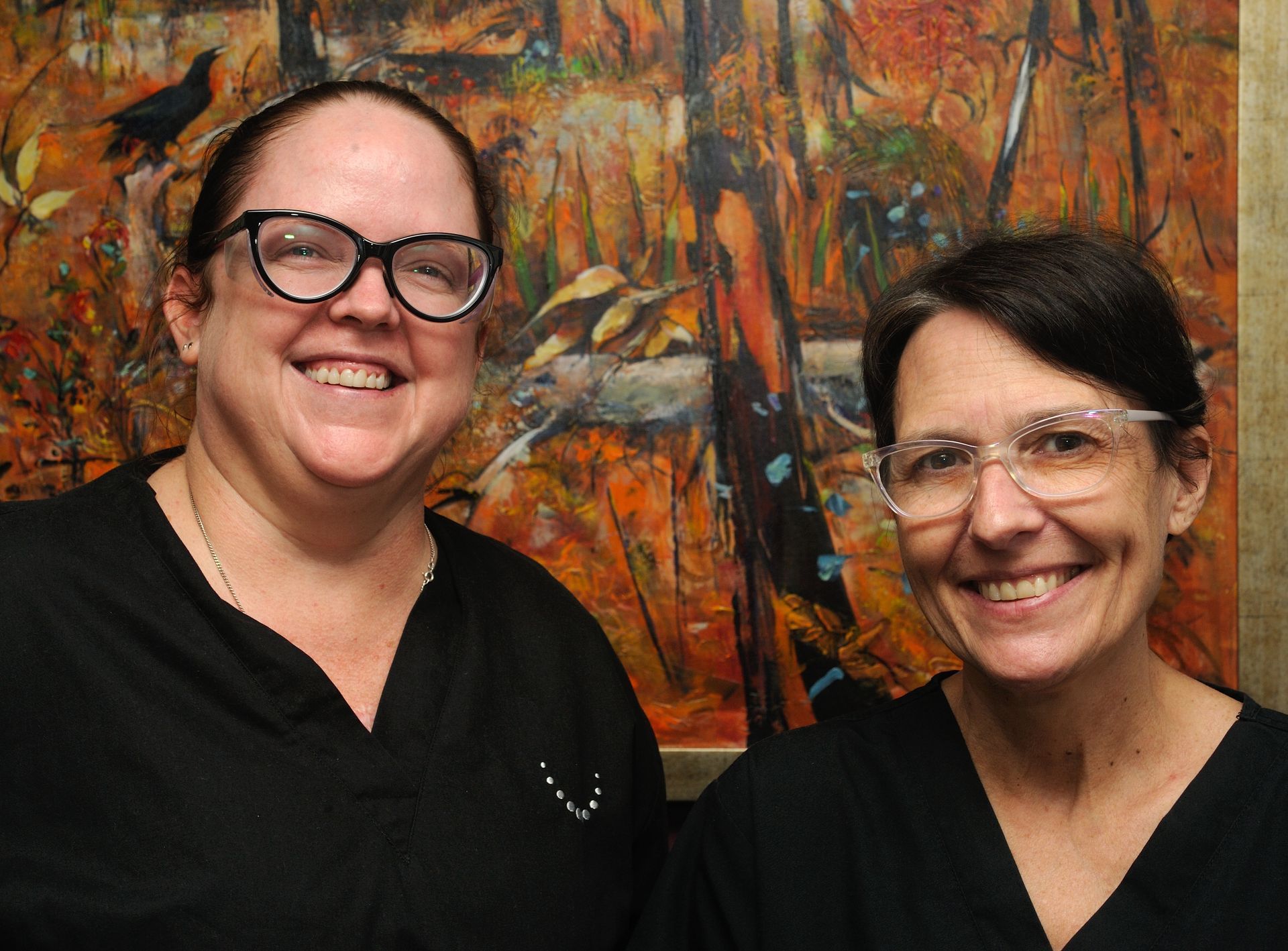 Two Women Wearing Glasses and Scrubs Are Smiling in Front of a Painting — Parap Family Dental in Parap, NT