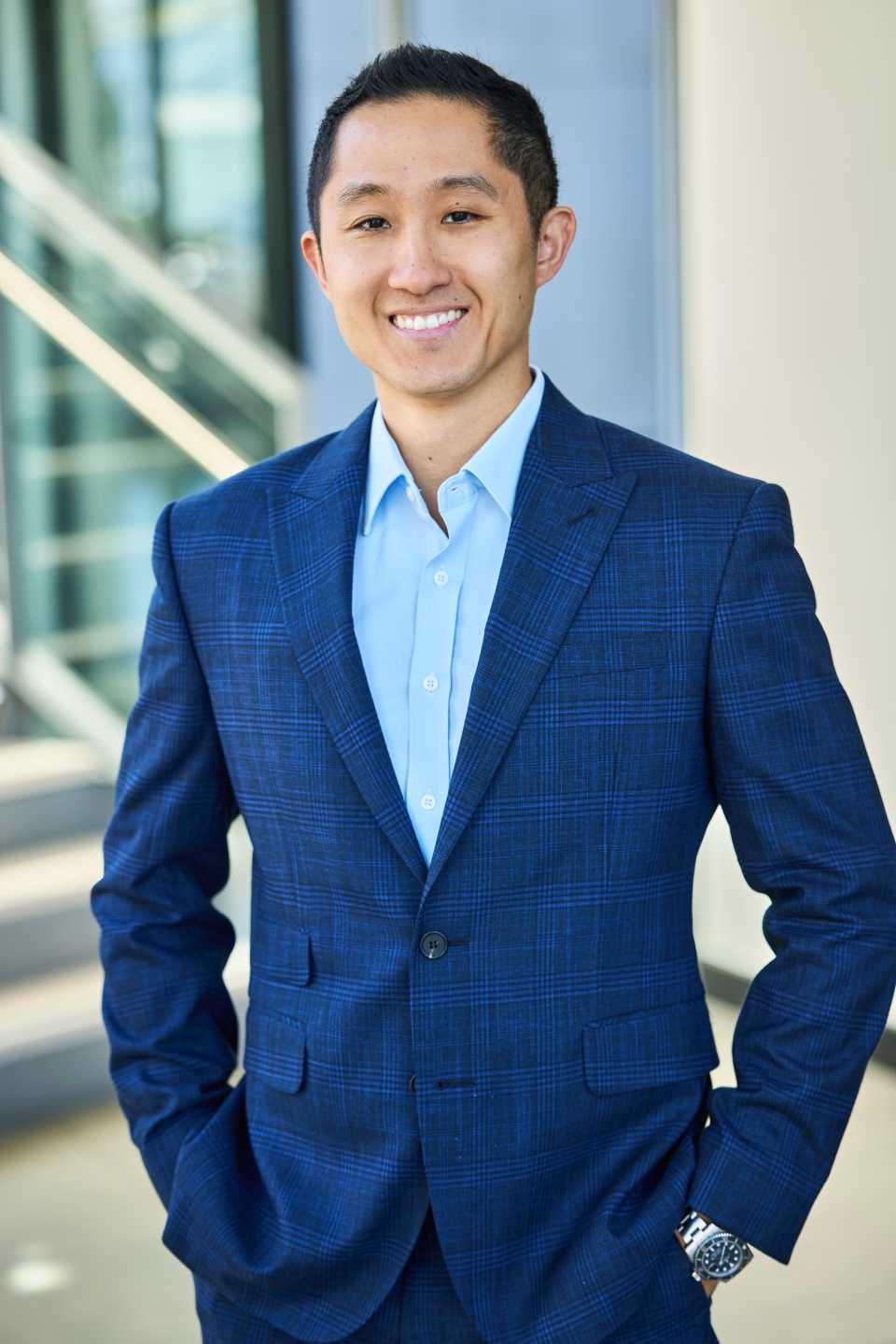Man in blue suit, smiling with hands in pockets, standing near building.