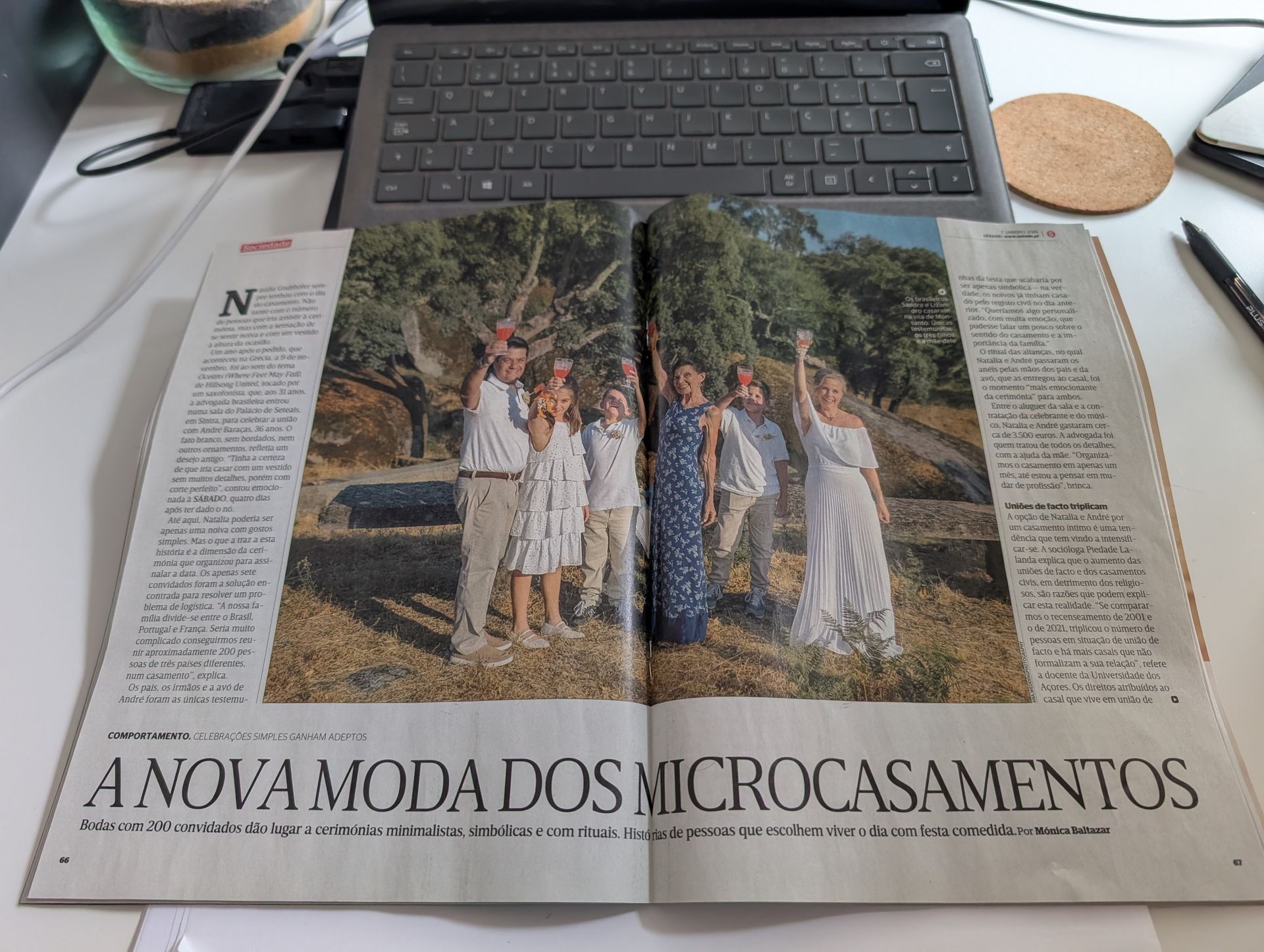 A professional photograph from a feature in Revista Sábado showing a family of five celebrating a bespoke vow renewal in the historic village of Monsanto, Portugal. The group is pictured in an outdoor setting with ancient trees and large granite boulders, holding glasses for a toast. The image represents a micro-wedding curated by Elope in Portugal.