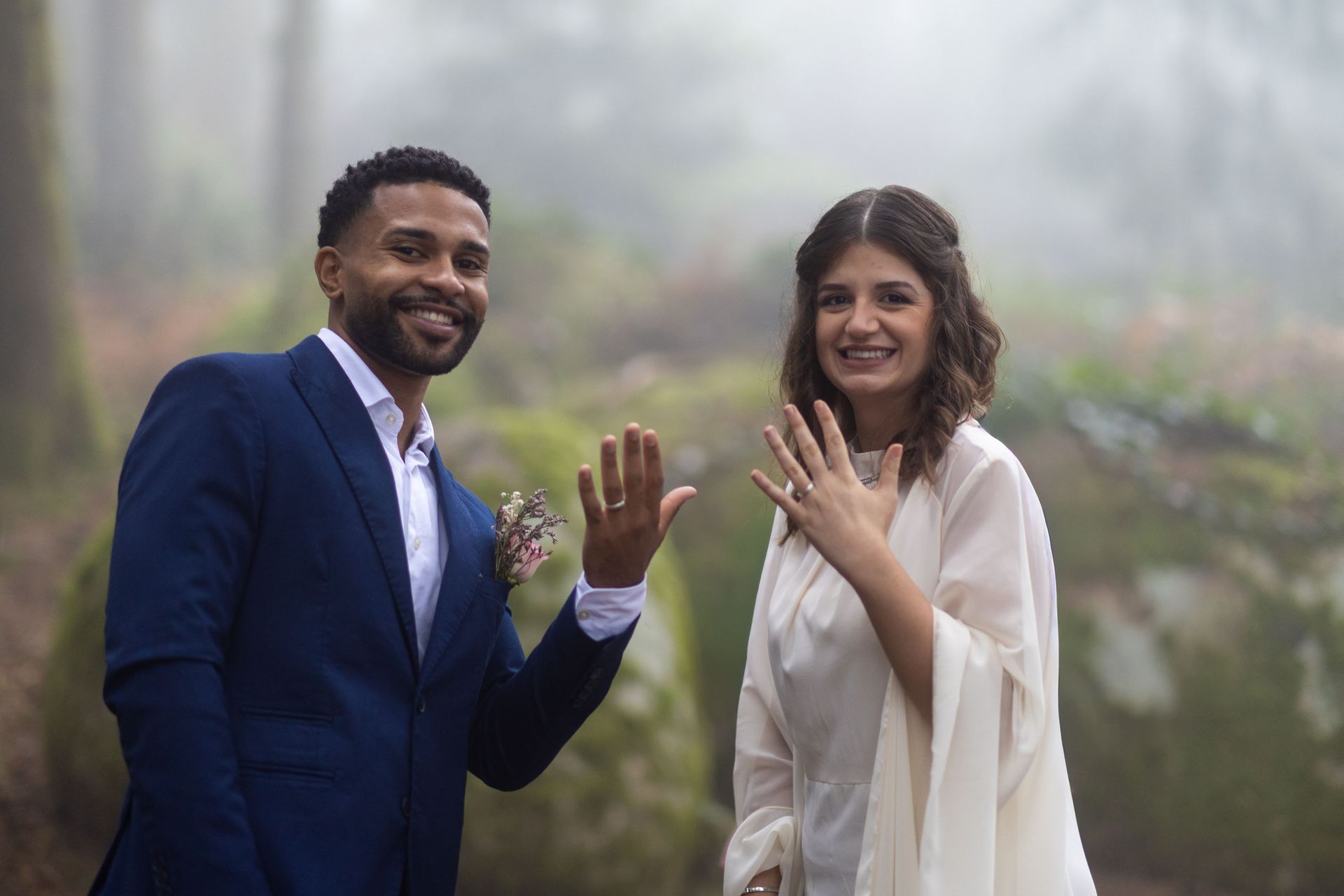 A close-up of the newly married couple in a foggy forest, smiling and showing off their wedding rings to the camera. The groom is in a blue suit and the bride in a white gown.