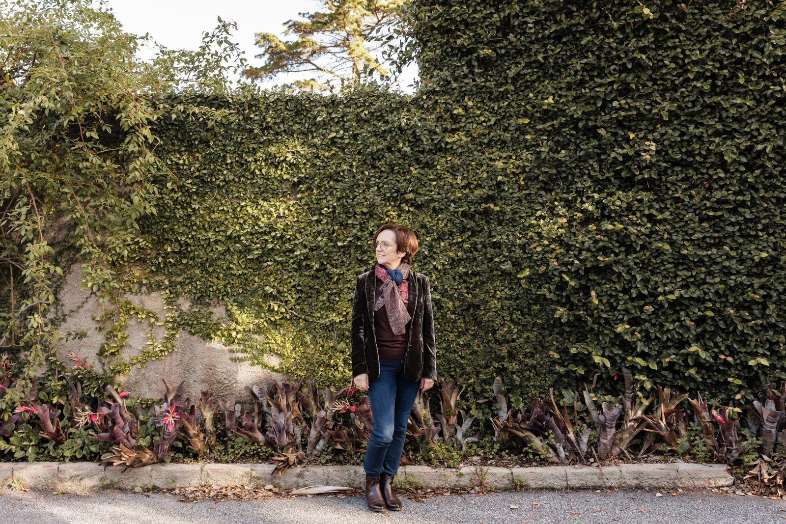 Anabela sits smiling on a large, mossy, felled tree log in a dense, foggy forest. She wears a cream-coloured cardigan over a patterned dress. The misty woods of Serra de Sintra create an ethereal, atmospheric backdrop.